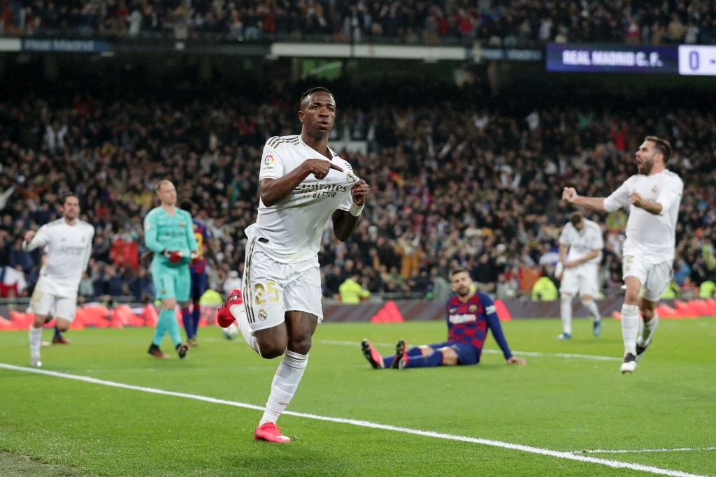 Vinicius se señala el escudo tras marcar el primer gol del partido y el inicio del dominio del Real Madrid. Por detrás, Piqué (hundido) en el suelo después de ver cómo el balón había rebotado en él para introducirse en la portería.